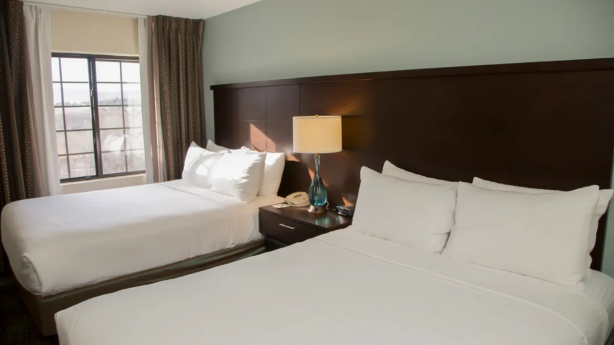 A serene bedroom featuring two double beds dressed in plush bedding, complemented by a cozy lamp and a large window offering natural light. The tranquil setting is designed for relaxation and comfort, with soft colors and inviting textures that speak to restorative sleep and winding down after an active day.