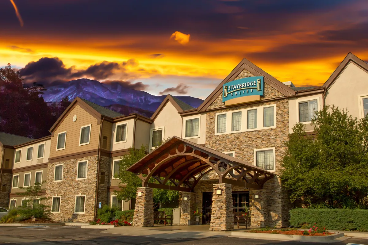 This image showcases the exterior of the property, featuring a striking stone facade, a warm wooden canopy, a clear hotel sign, and a beautifully landscaped garden with mountain peaks in the background. The scene suggests a welcoming atmosphere for families arriving for their Colorado Springs adventure, while the mountain backdrop hints at the nearby natural attractions.