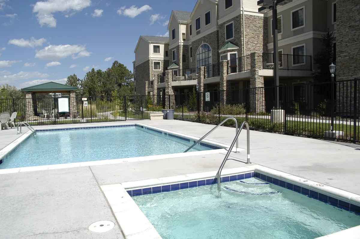 This image shows a spacious outdoor pool area with a swimming pool and a hot tub side by side, surrounded by lush greenery and the apartment building in the background. There are people relaxing in lounge chairs, enjoying the water and the sunshine, with a tranquil and leisurely atmosphere that invites relaxation and socialization.